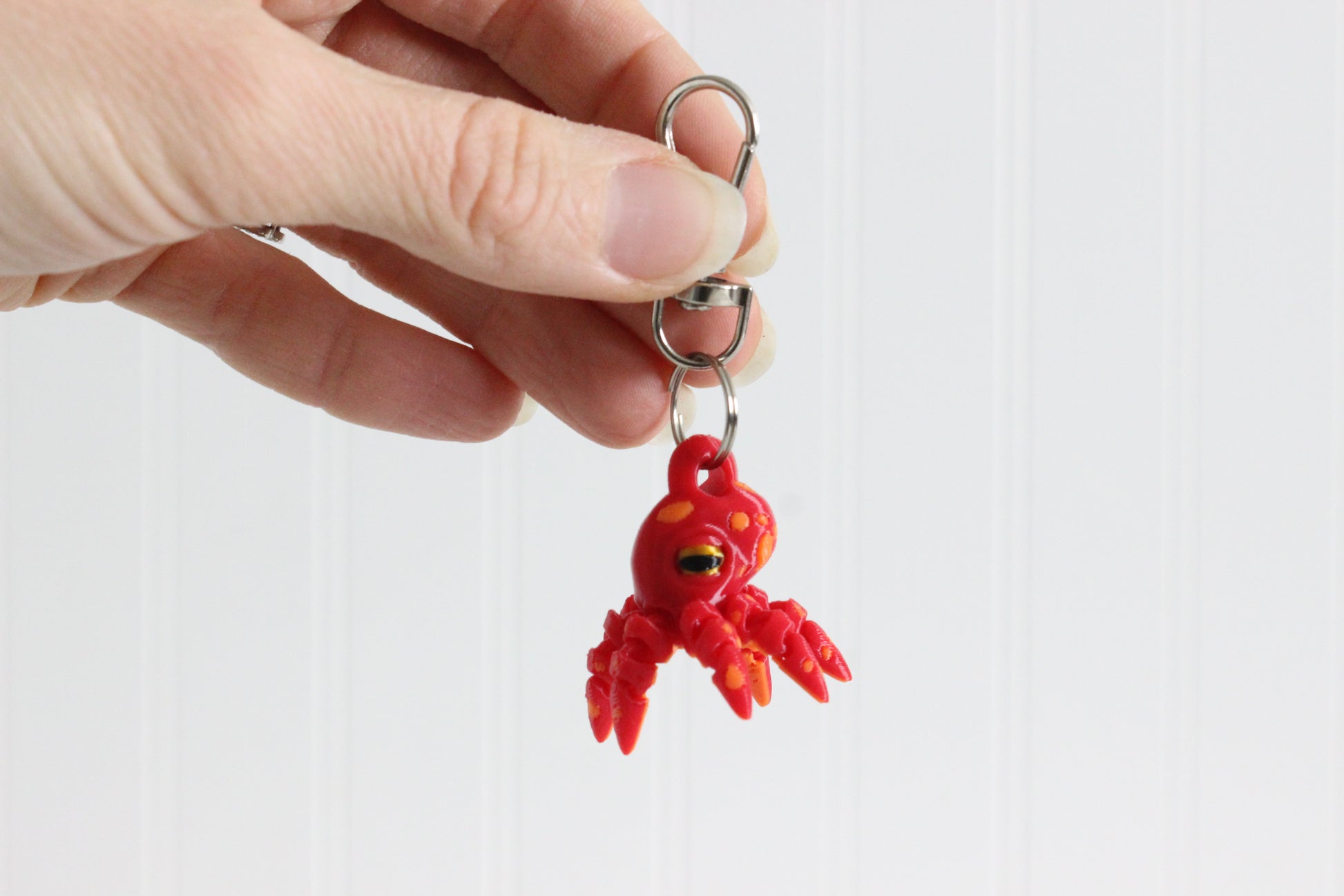Hand holding the 3D-printed octopus with curled tentacles keychain to show its size, colors, and silver swivel clip.