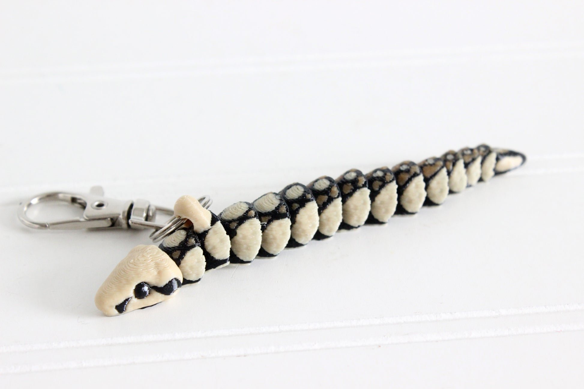 Articulated boa constrictor snake keychain with black and cream scale pattern, lying flat on a white surface with swivel quick-clip key ring