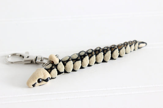 Articulated boa constrictor snake keychain with black and cream scale pattern, lying flat on a white surface with swivel quick-clip key ring