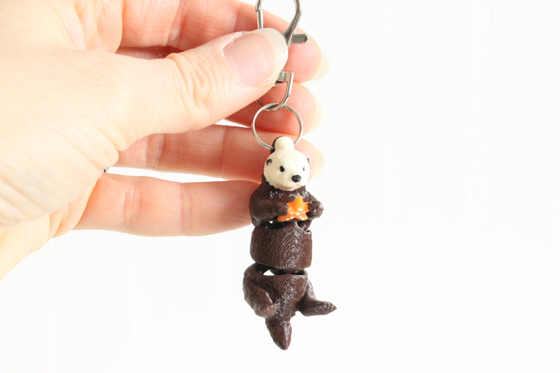 Hand holding 3D-printed brown sea otter keychain to show its size, colors, and silver swivel clip.