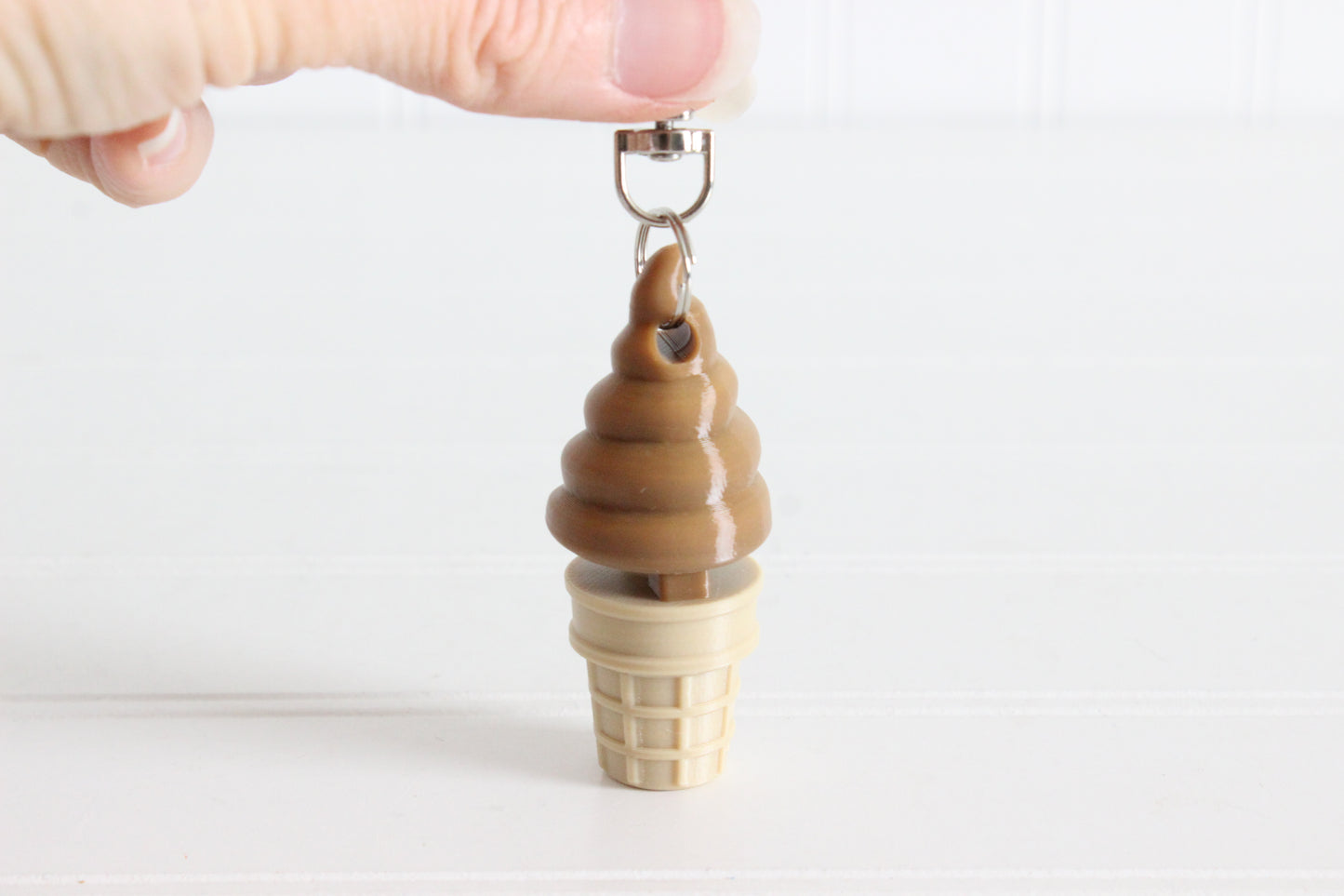 Hand holding a chocolate soft-serve ice cream cone keychain to show its size.