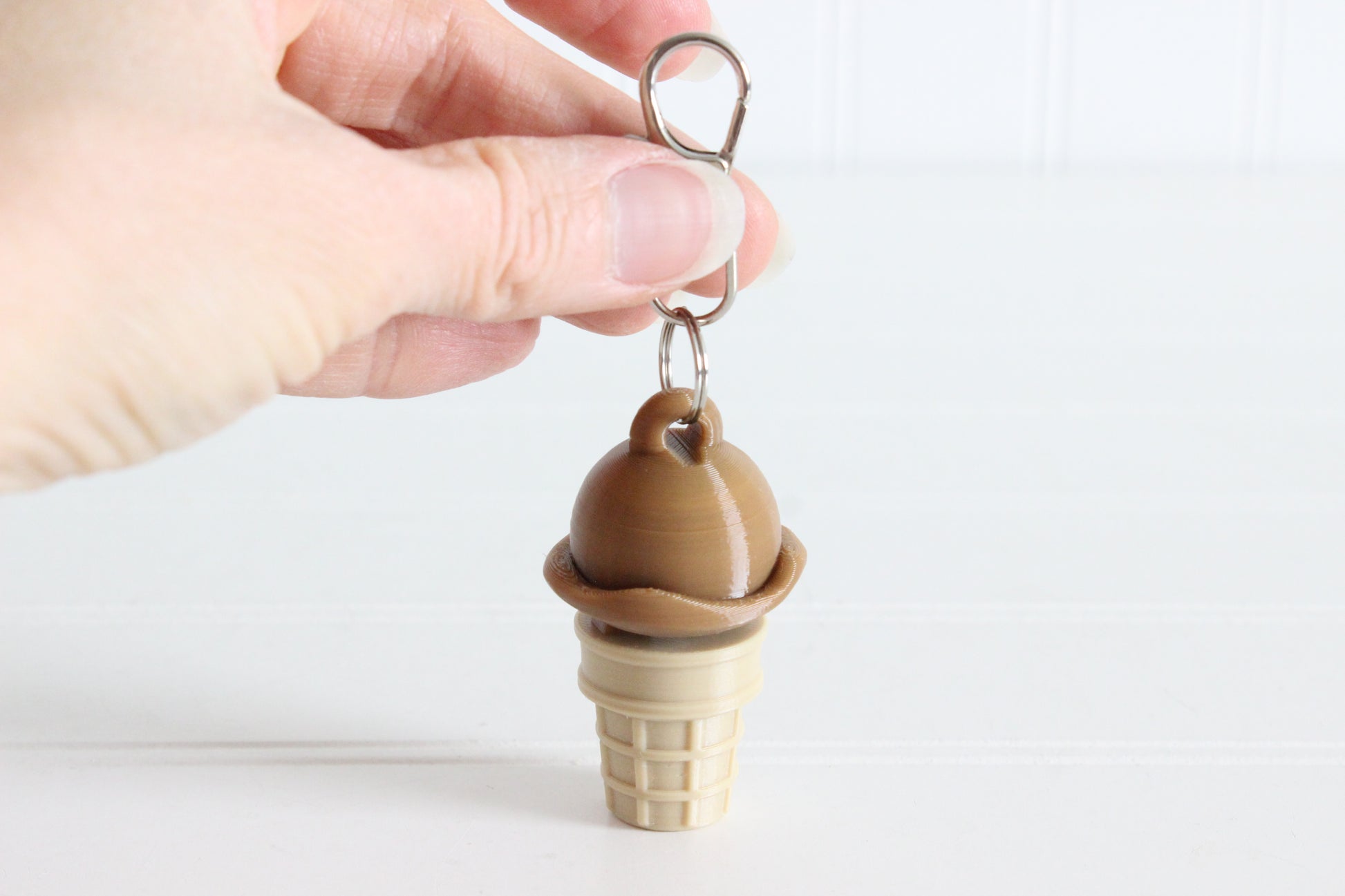 Hand holding 3D-printed chocolate ice cream cone keychain to show its size, colors, and silver swivel clip.