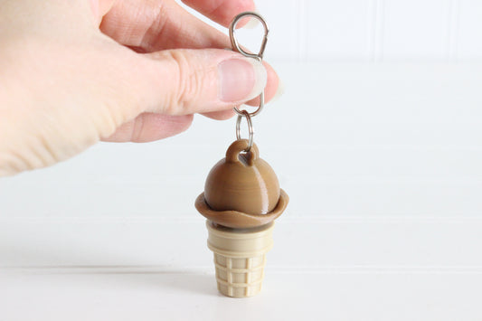 Hand holding 3D-printed chocolate ice cream cone keychain to show its size, colors, and silver swivel clip.
