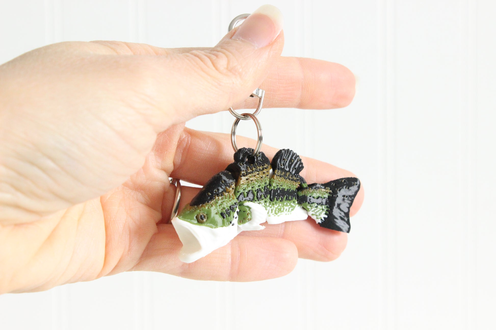 Hand holding the 3D-printed green largemouth bass keychain to show its size, colors, and silver swivel clip.