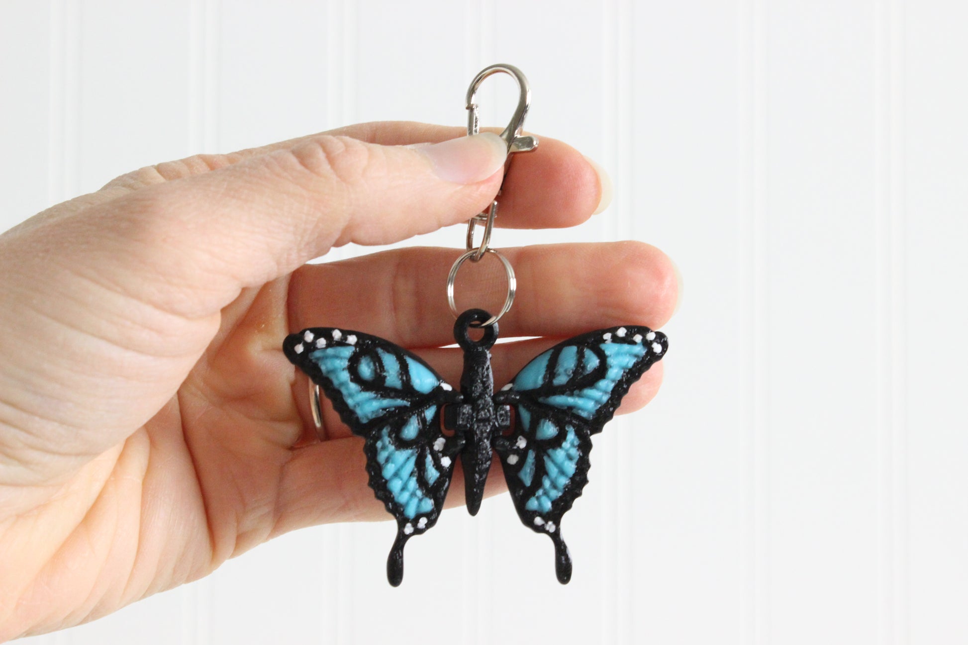 Hand holding 3D-printed blue butterfly keychain to show its size, colors, and silver swivel clip.