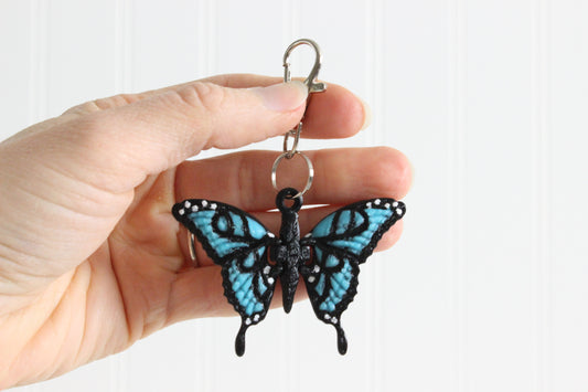 Hand holding 3D-printed blue butterfly keychain to show its size, colors, and silver swivel clip.