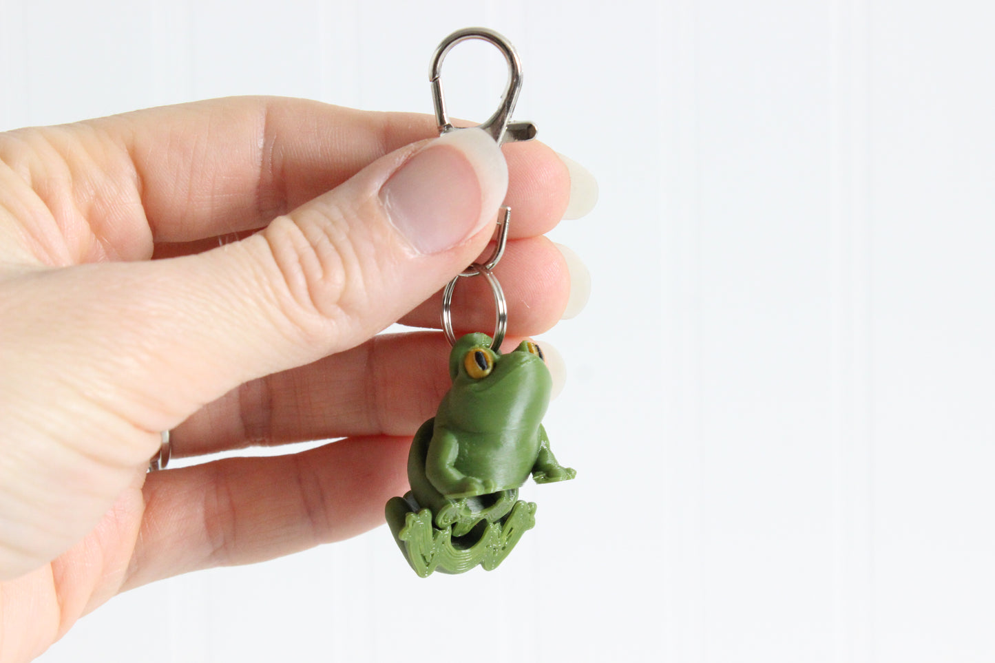 Hand holding the 3D-printed green frog keychain to show its size, colors, and silver swivel clip.