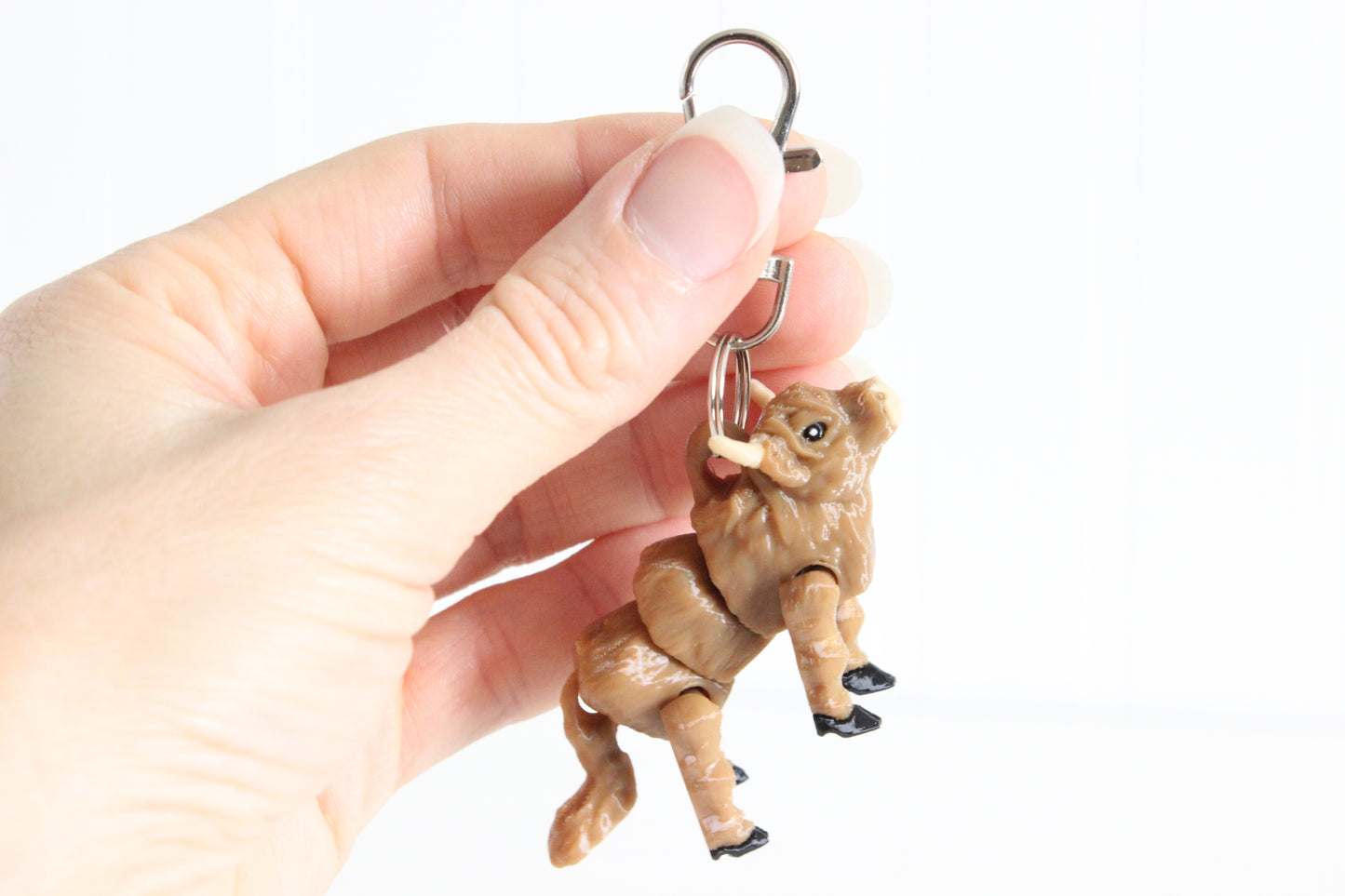 Hand holding the 3D-printed shaggy brown Highland cow keychain to show its size, colors, and silver swivel clip.