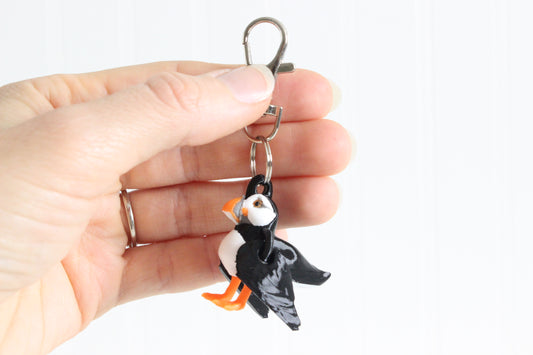 Hand holding 3D-printed black, white, and orange puffin keychain to show its size, colors, and silver swivel clip.