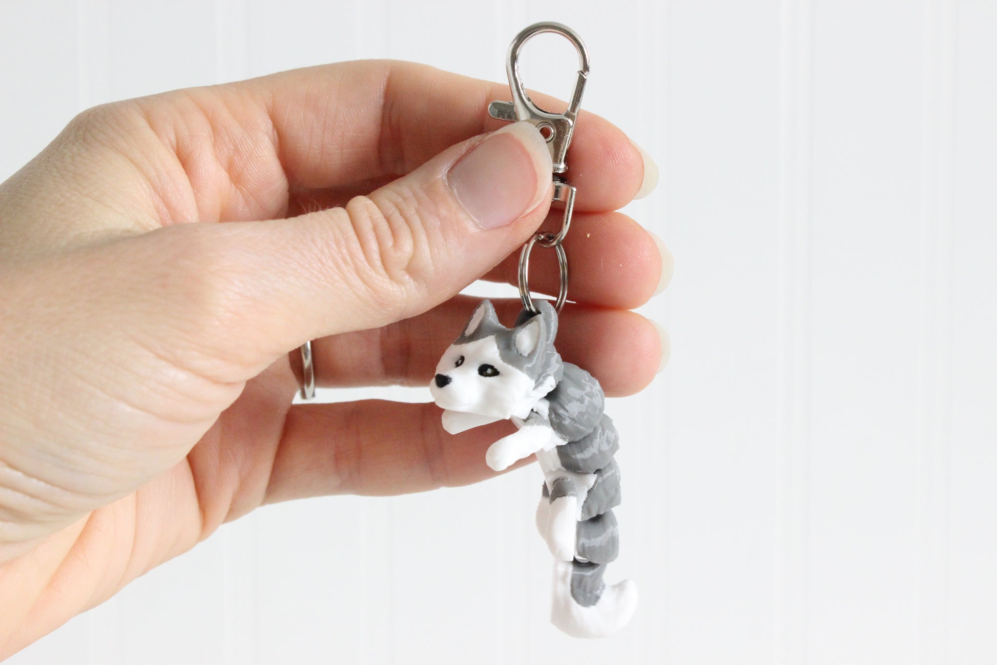 Hand holding the 3D-printed gray-and-white husky keychain to show its size, colors, and silver swivel clip.