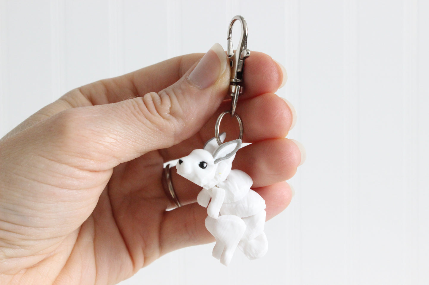 Hand holding the 3D-printed white snow rabbit keychain to show its size, colors, and silver swivel clip.