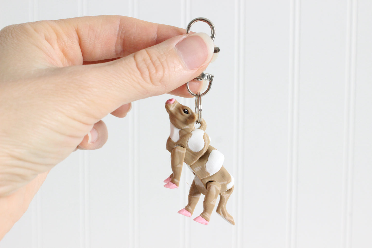 Hand holding the 3D-printed brown-and-white cow keychain to show its size, colors, and silver swivel clip.