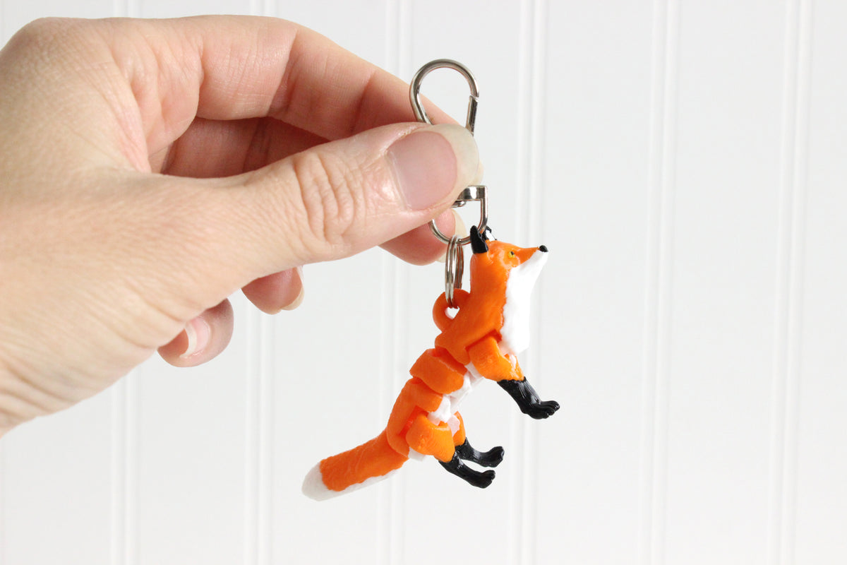 Hand holding the 3D-printed orange-red fox keychain to show its size, colors, and silver swivel clip.